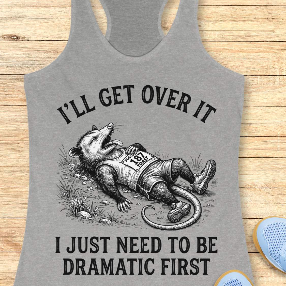 A heather gray racerback tank top laid flat on a light wood surface, featuring a black graphic of an opossum lying down with the text 'I'LL GET OVER IT' above and 'I JUST NEED TO BE DRAMATIC FIRST' below.