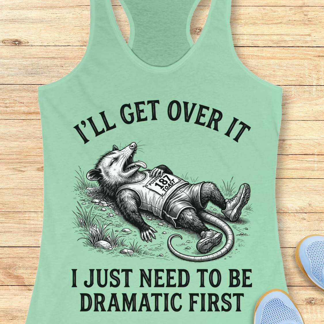 A mint green racerback tank top laid flat on a light wood background, featuring a humorous graphic of a raccoon dressed as a racehorse with the text 'I'LL GET OVER IT I JUST NEED TO BE DRAMATIC FIRST'.