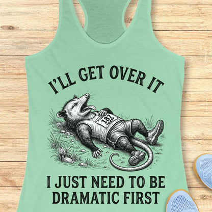 A mint green racerback tank top laid flat on a light wood background, featuring a humorous graphic of a raccoon dressed as a racehorse with the text 'I'LL GET OVER IT I JUST NEED TO BE DRAMATIC FIRST'.