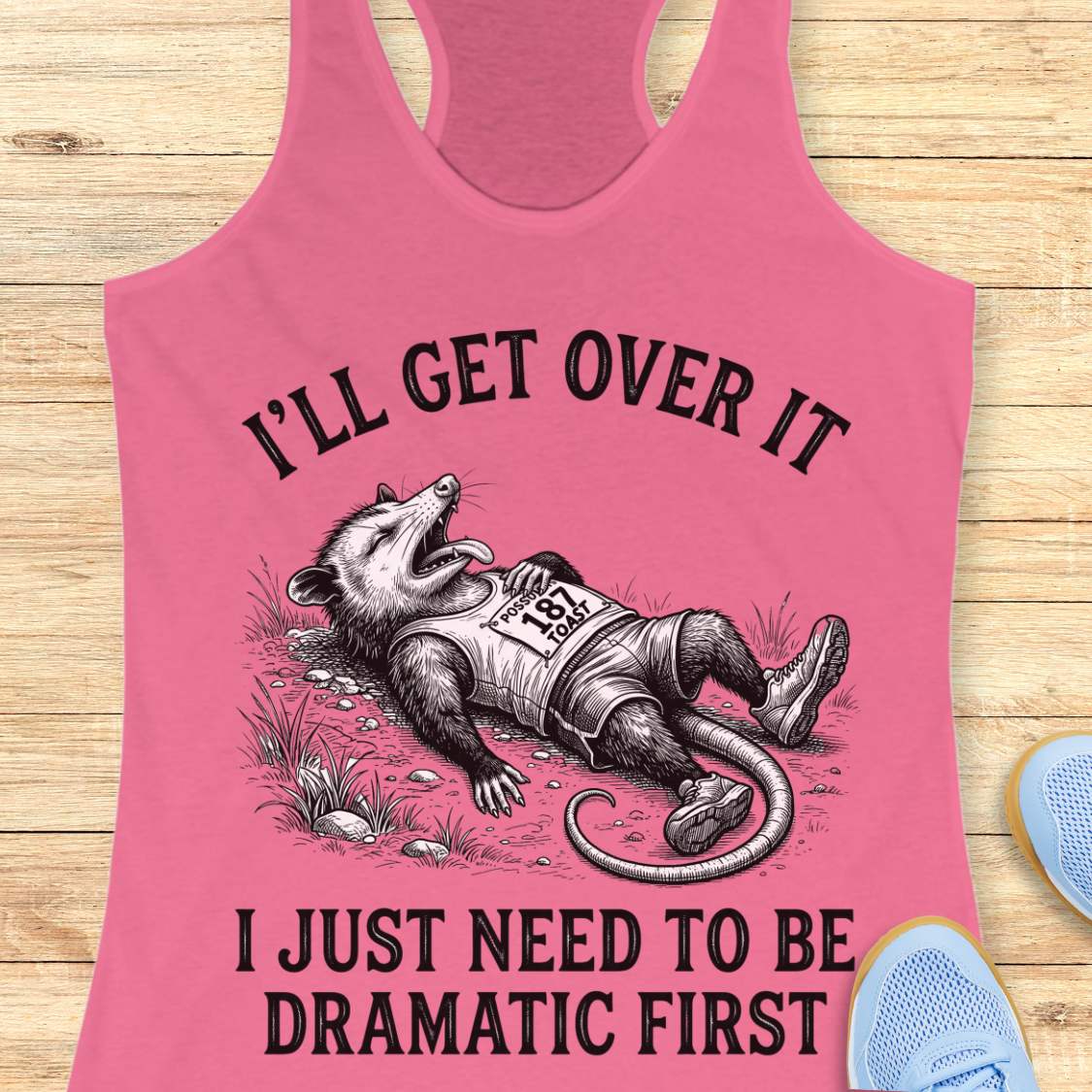A pink racerback tank top laid flat on a light wood surface, featuring a black graphic of a raccoon wearing a race bib and the text 'I'LL GET OVER IT I JUST NEED TO BE DRAMATIC FIRST.' Partial blue shoes are visible at the lower right as styling props.