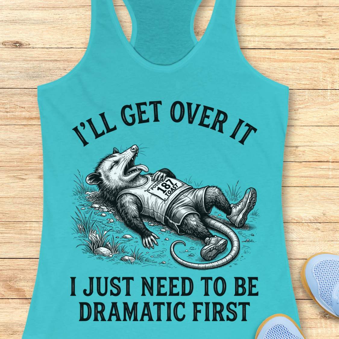 A turquoise racerback tank top laid flat on a light wood floor, featuring a humorous graphic of a raccoon lying down with the text 'I’LL GET OVER IT I JUST NEED TO BE DRAMATIC FIRST'.