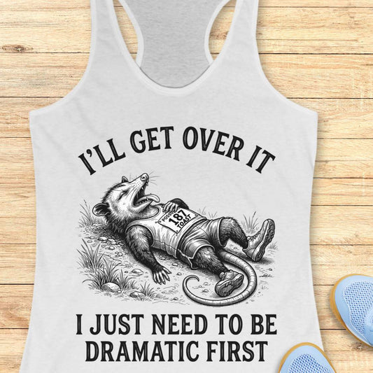 A white racerback tank top laid flat on a light wood background, featuring a black graphic of an opossum lying down with the text 'I'LL GET OVER IT' above and 'I JUST NEED TO BE DRAMATIC FIRST' below. Partial blue shoes are visible at the bottom right as styling props.