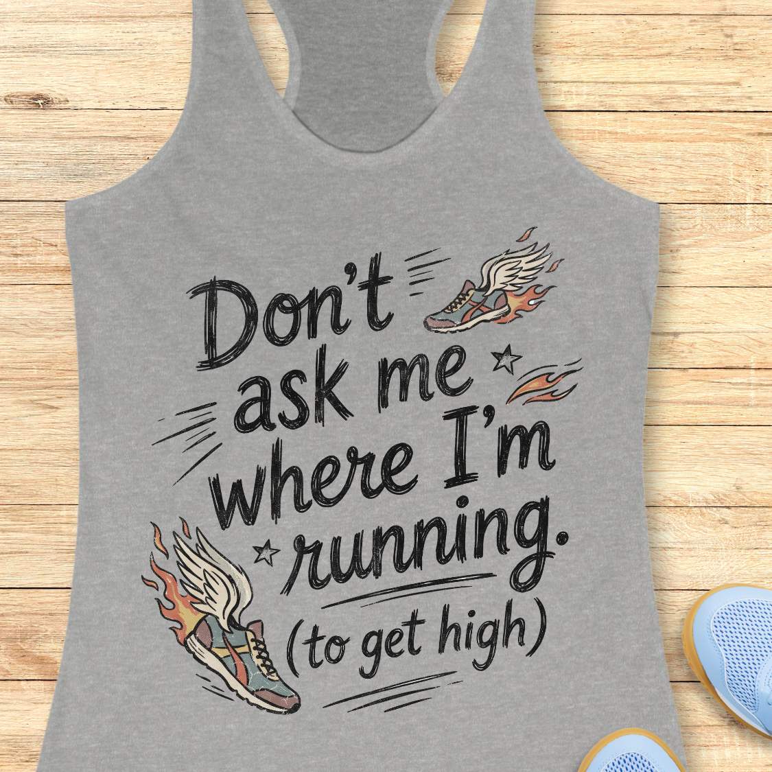 A heather gray racerback tank top laid flat on a light wood surface, featuring a black hand-lettered graphic that reads 'Don't ask me where I'm running. (to get high)' with two illustrated winged running shoes in orange, gray, and white.