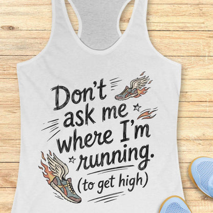 A white racerback tank top laid flat on a light wood background, printed with the phrase “Don’t ask me where I’m running. (to get high)” in black script, accompanied by two illustrated winged running shoes in orange, brown, and gray.