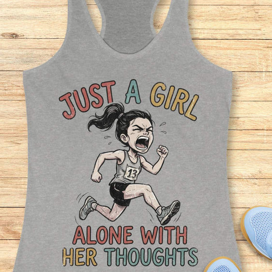 A heather gray racerback tank top laid flat on a light wood background, featuring a graphic of a running girl with the text 'JUST A GIRL ALONE WITH HER THOUGHTS' in multicolored lettering.