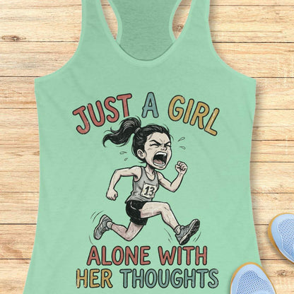 A mint green racerback tank top laid flat on a light wood background, featuring a cartoon illustration of a running girl with the text 'JUST A GIRL ALONE WITH HER THOUGHTS' in multicolored lettering.