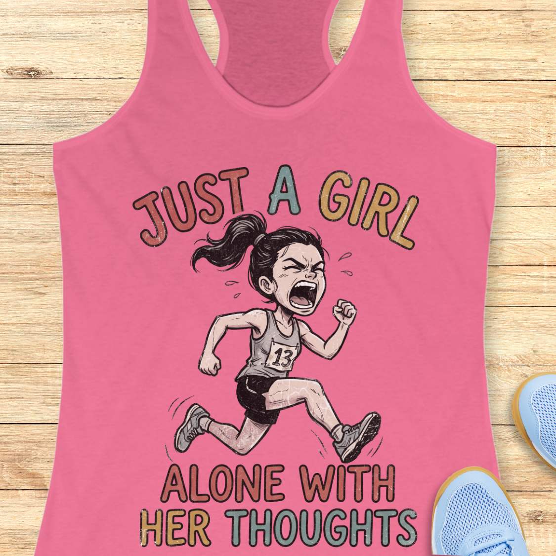 A pink racerback tank top laid flat on a light wood surface, featuring a cartoon illustration of a running girl and the text 'JUST A GIRL ALONE WITH HER THOUGHTS.' Partial blue shoes are visible at the edges as styling props.