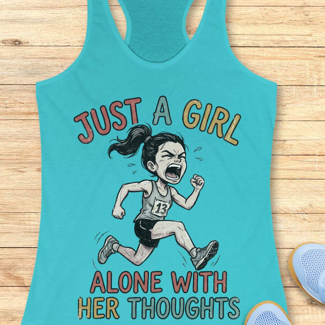 A turquoise racerback tank top laid flat on a light wood surface, featuring a cartoon illustration of a running girl with the text 'JUST A GIRL ALONE WITH HER THOUGHTS' in multicolored lettering.