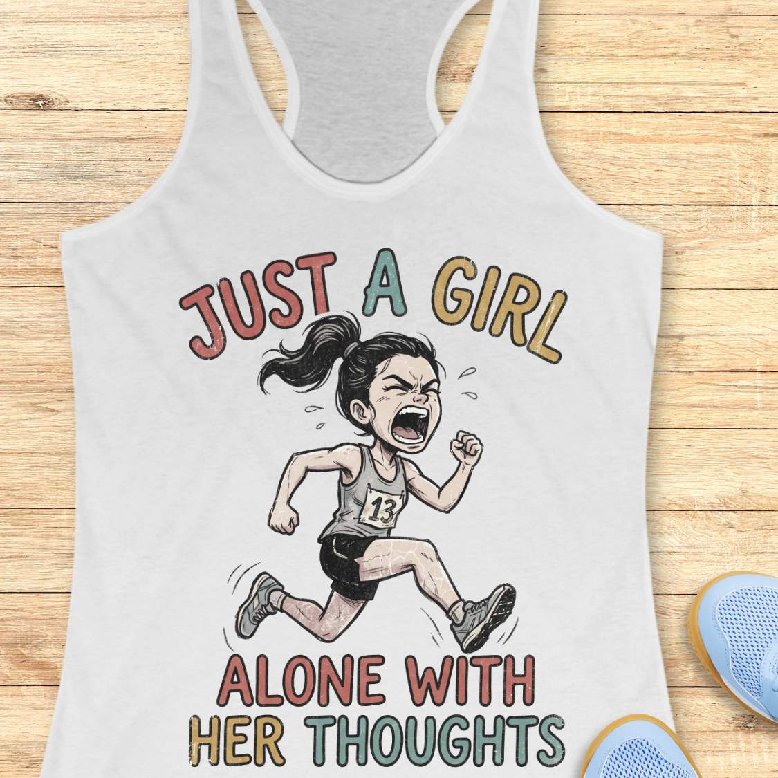 A white racerback tank top laid flat on a light wood background, featuring a cartoon illustration of a running girl and the text 'JUST A GIRL ALONE WITH HER THOUGHTS' in multicolored lettering. Partial blue shoes are visible at the bottom right as styling props.