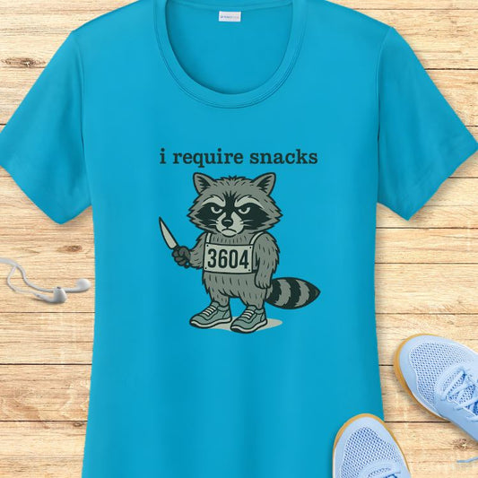 Teal t-shirt featuring a cartoon grey raccoon wearing a "3604" bib, holding a small knife, and the text "i require snacks", presented on a wooden surface with light blue running shoes and white earbuds