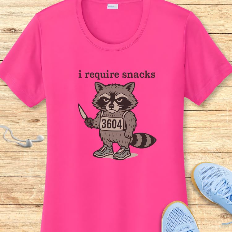 Photograph of a hot pink t-shirt featuring a cartoon raccoon holding a knife and wearing a '3604' race bib, with 'i require snacks' text above, displayed on a wooden surface alongside blue athletic shoes and white earbuds