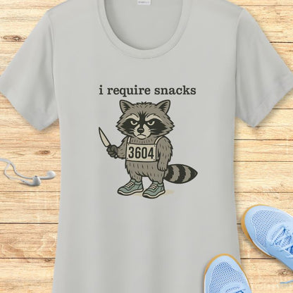 Digital print on a grey t-shirt featuring a cartoon raccoon wearing a '3604' bib and blue sneakers, holding a knife, with the text 'i require snacks' above, displayed on a wooden surface with white earbuds and a blue running shoe