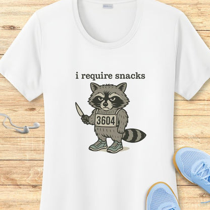White cotton t-shirt featuring a digital print of an angry cartoon raccoon holding a knife, wearing blue sneakers and a "3604" tag, with the text i require snacks above it, displayed on a light wooden surface beside white earbuds and light blue athletic shoes