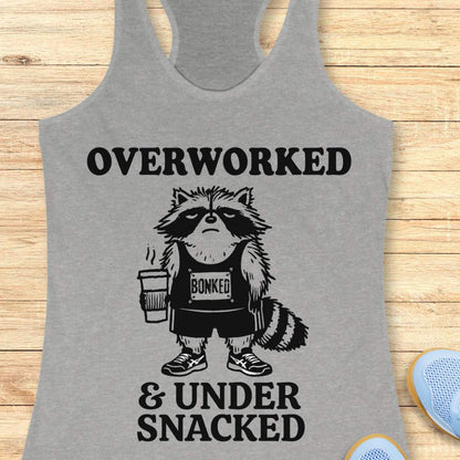A heather gray racerback tank top laid flat on a light wood surface, featuring a black graphic of a raccoon wearing a 'Bonked' tank top and shorts while holding a coffee cup. The text reads 'OVERWORKED & UNDER SNACKED'.
