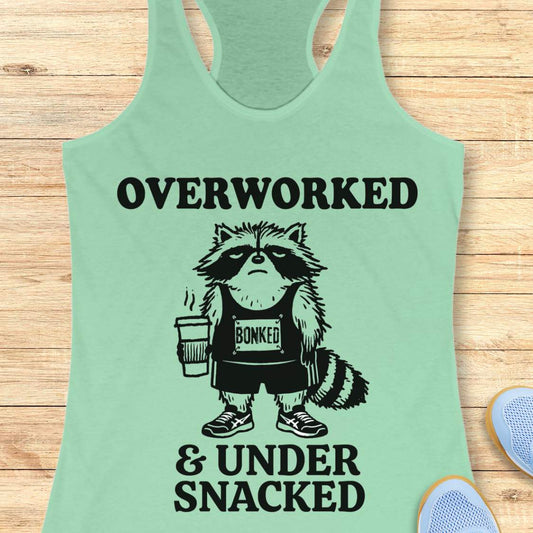 A mint green racerback tank top laid flat on a light wood background, featuring a black graphic of a raccoon holding a coffee cup with the text 'OVERWORKED & UNDER SNACKED'.