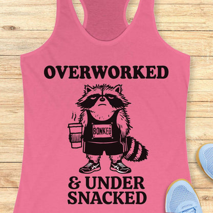 A pink racerback tank top laid flat on a light wood surface, featuring a black graphic of a raccoon wearing a 'BONKED' shirt and holding a coffee cup, with the text 'OVERWORKED & UNDER SNACKED'.