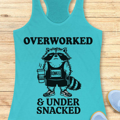 A turquoise racerback tank top laid flat on a light wood surface, featuring a black graphic of a raccoon wearing a 'BONKED' tank top and holding a coffee cup, with the text 'OVERWORKED & UNDER SNACKED'.