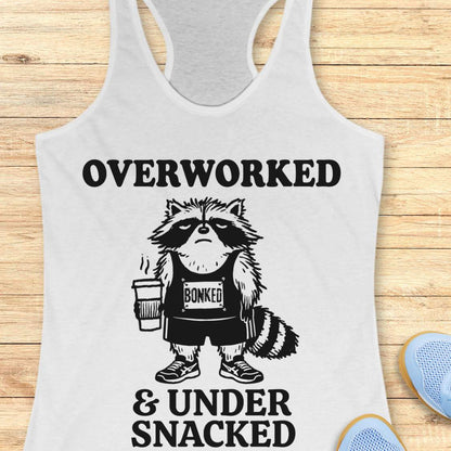 A white racerback tank top laid flat on a light wood background, featuring a black graphic of a raccoon wearing a 'Bonked' shirt and holding a coffee cup, with the text 'OVERWORKED & UNDER SNACKED'.