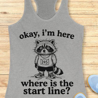 A heather gray racerback tank top laid flat on a light wood surface, featuring a black graphic of a raccoon wearing a race bib numbered 245, with the text 'okay, i'm here where is the start line?'.