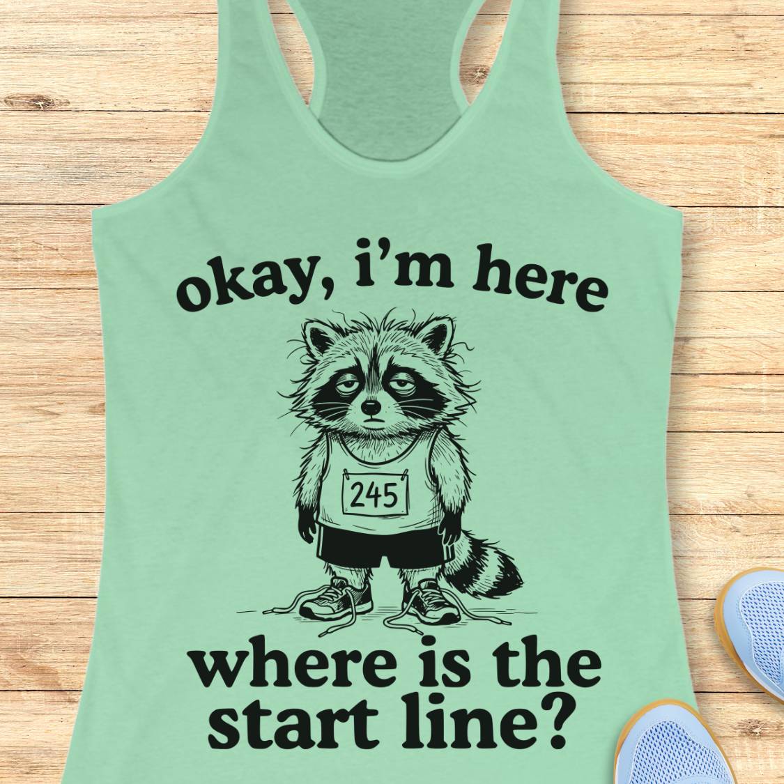 A mint green racerback tank top laid flat on a light wood background, featuring a black graphic of a raccoon wearing a race bib and the text 'okay, i'm here where is the start line?'. Partial blue shoes are visible at the bottom right as styling props.