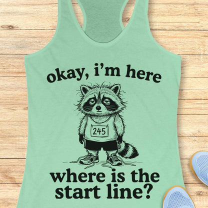 A mint green racerback tank top laid flat on a light wood background, featuring a black graphic of a raccoon wearing a race bib and the text 'okay, i'm here where is the start line?'. Partial blue shoes are visible at the bottom right as styling props.
