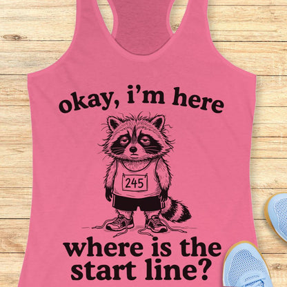 A pink racerback tank top laid flat on a light wood surface, featuring a black graphic of a raccoon wearing a race bib and the text 'okay, i'm here where is the start line?'. Partial blue sneakers are visible at the edges as styling props.