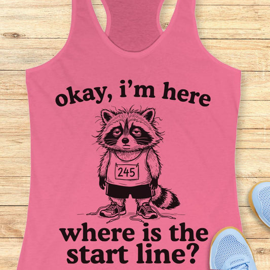A pink racerback tank top laid flat on a light wood surface, featuring a black graphic of a raccoon wearing a race bib and the text 'okay, i'm here where is the start line?'. Partial blue sneakers are visible at the edges as styling props.