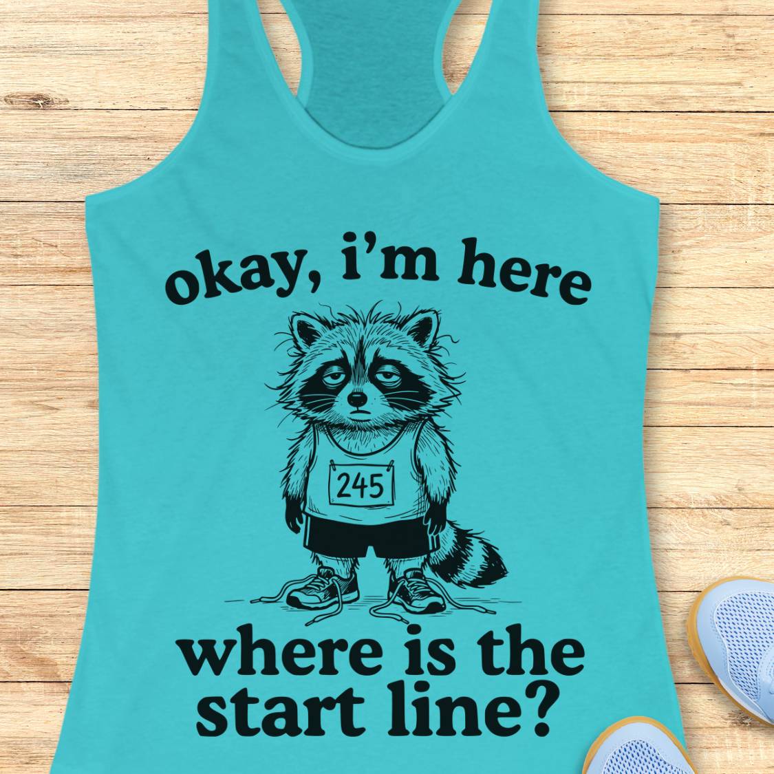 A turquoise racerback tank top laid flat on a light wood surface, featuring a black graphic of a raccoon wearing a race bib and the text 'okay, i'm here where is the start line?'. Part of a white shoe is visible at the bottom right.