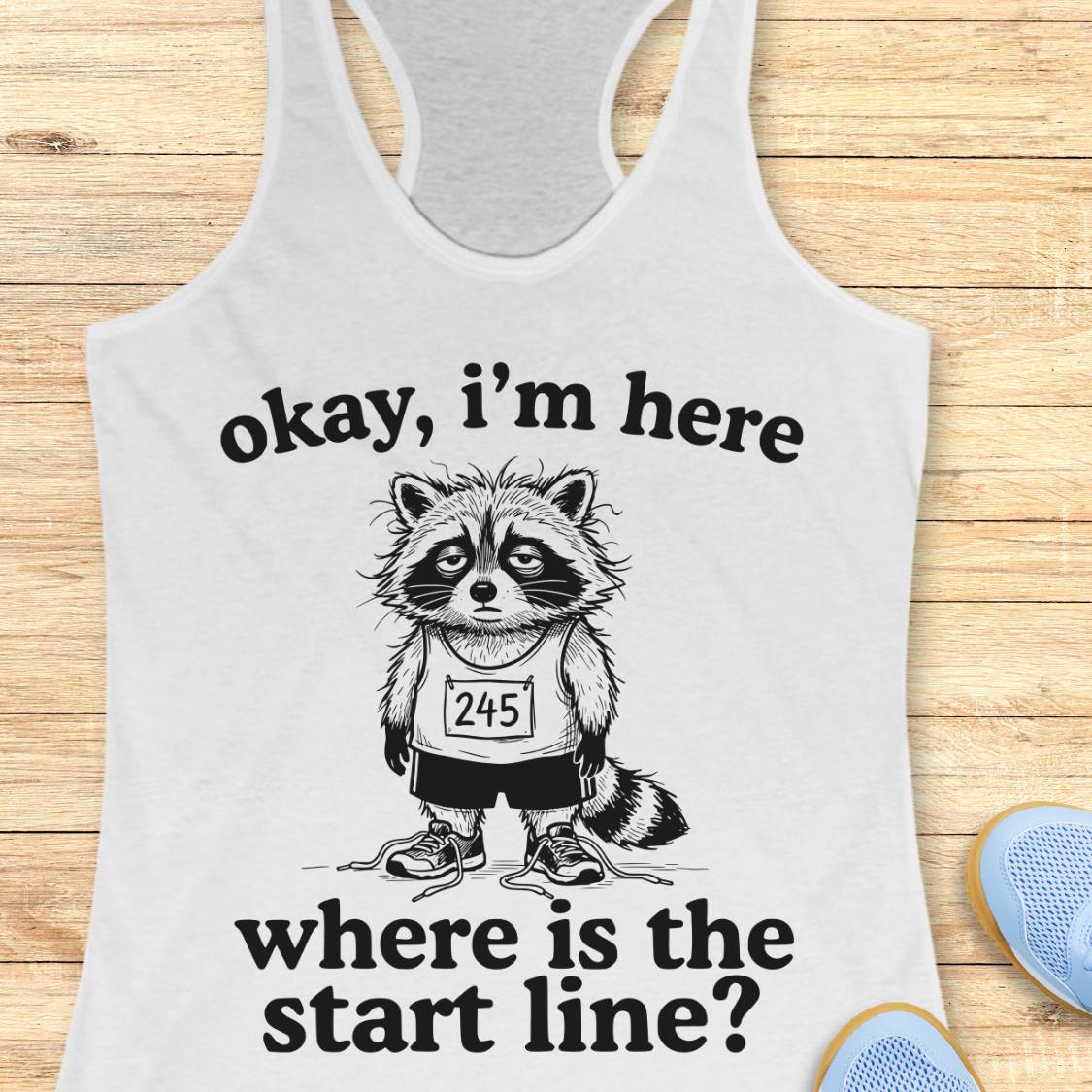 A white racerback tank top laid flat on a light wood surface, featuring a black graphic of a raccoon wearing a race bib numbered 245, with the text 'okay, i'm here where is the start line?'. Partial blue shoes are visible at the bottom right as styling props.