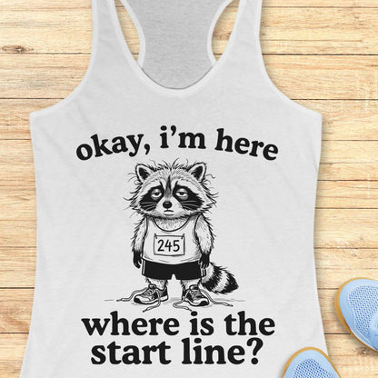 A white racerback tank top laid flat on a light wood surface, featuring a black graphic of a raccoon wearing a race bib numbered 245, with the text 'okay, i'm here where is the start line?'. Partial blue shoes are visible at the bottom right as styling props.