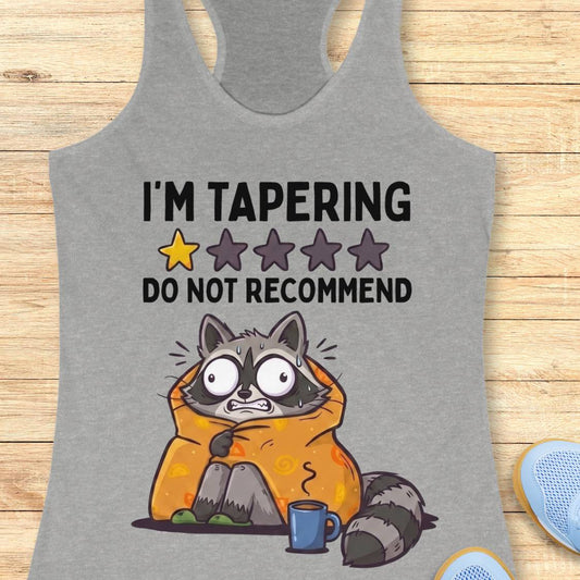 A heather gray racerback tank top laid flat on a light wood background, featuring a humorous graphic of a raccoon wrapped in an orange blanket with the text 'I'M TAPERING DO NOT RECOMMEND' and a star rating graphic.
