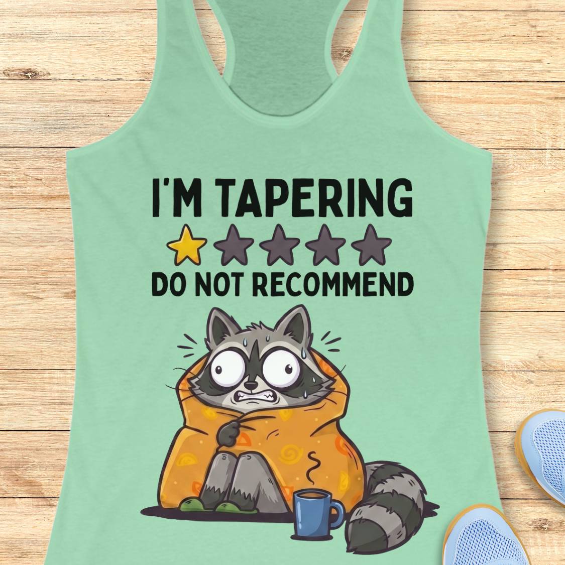 A mint green racerback tank top laid flat on a light wood background, featuring a humorous graphic of a raccoon wrapped in an orange blanket with a blue mug and the text 'I'M TAPERING DO NOT RECOMMEND' with a yellow and gray star rating.