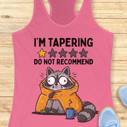 A pink racerback tank top laid flat on a light wood surface, featuring a humorous graphic of a raccoon wrapped in an orange blanket with the text 'I'M TAPERING DO NOT RECOMMEND' and a one-star review graphic. Partial blue sneakers are visible at the bottom right as styling props.