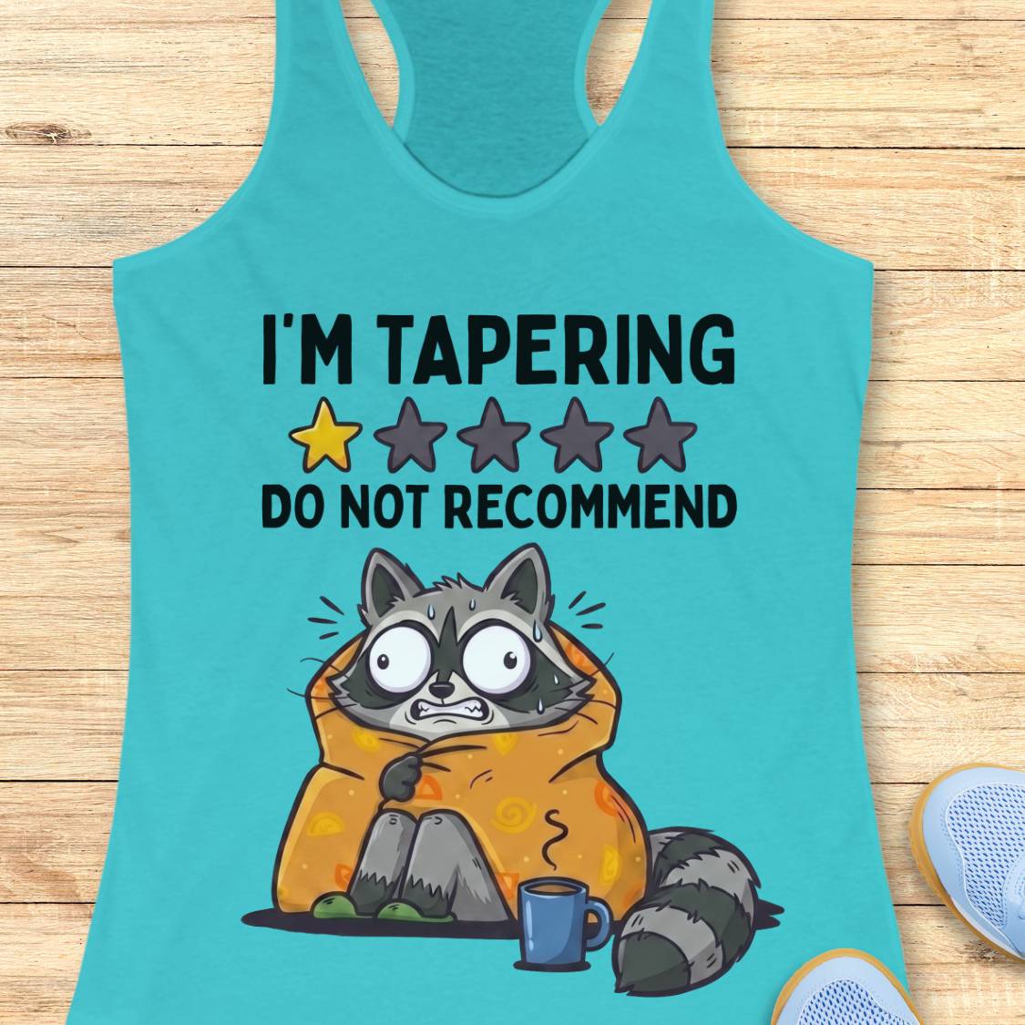 A turquoise racerback tank top laid flat on a light wood background, featuring a humorous graphic of a raccoon wrapped in an orange blanket with a coffee mug and the text 'I'M TAPERING DO NOT RECOMMEND' with a one-star review graphic.
