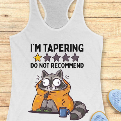 A white racerback tank top laid flat on a light wood background, featuring a cartoon raccoon wrapped in an orange blanket with the text 'I'M TAPERING DO NOT RECOMMEND' and a star rating graphic.
