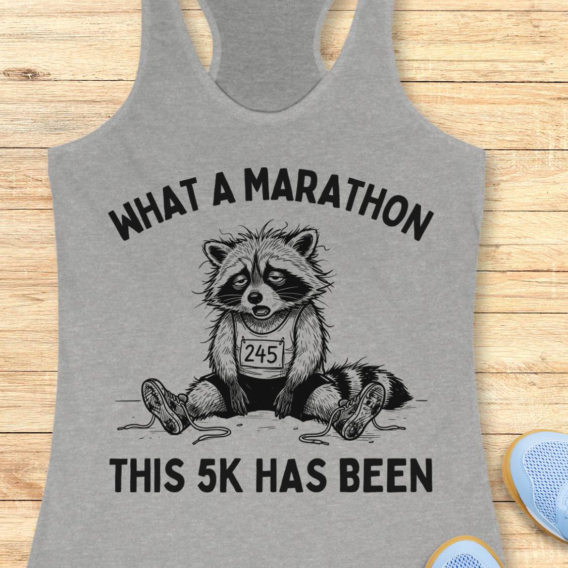 A heather gray racerback tank top laid flat on a light wood surface, featuring a black graphic of a raccoon wearing a race bib with the text 'WHAT A MARATHON THIS 5K HAS BEEN'.