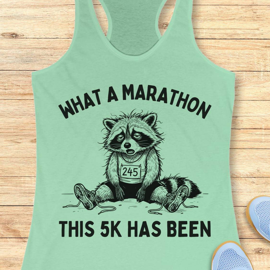 A mint green racerback tank top laid flat on a light wood background, featuring a black graphic of a raccoon wearing a race bib with the number 245 and the text 'WHAT A MARATHON THIS 5K HAS BEEN'.