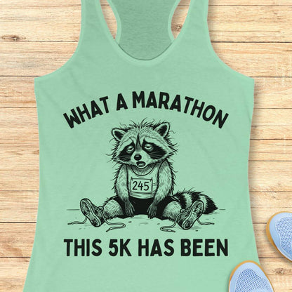A mint green racerback tank top laid flat on a light wood background, featuring a black graphic of a raccoon wearing a race bib with the number 245 and the text 'WHAT A MARATHON THIS 5K HAS BEEN'.