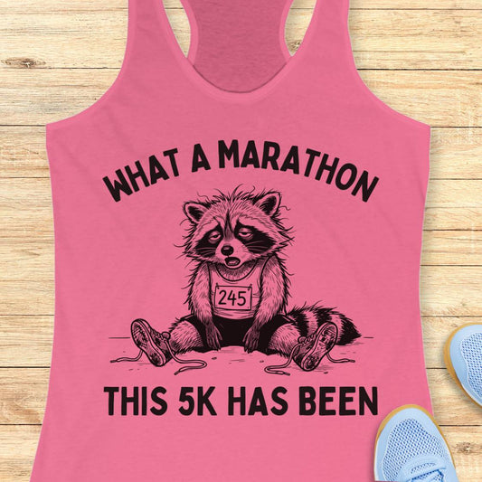 A pink racerback tank top laid flat on a light wood surface, featuring a black graphic of a raccoon wearing a race bib and the text 'WHAT A MARATHON THIS 5K HAS BEEN.' Partial blue shoes are visible at the lower right as styling props.