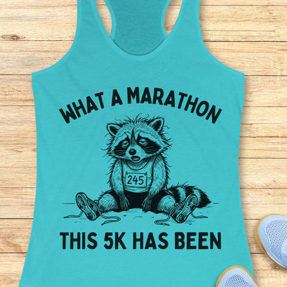 A turquoise racerback tank top laid flat on a light wood surface, featuring a black graphic of a sad raccoon wearing a race bib that says 245, with the text 'WHAT A MARATHON THIS 5K HAS BEEN'. A partial white sneaker is visible at the bottom right.