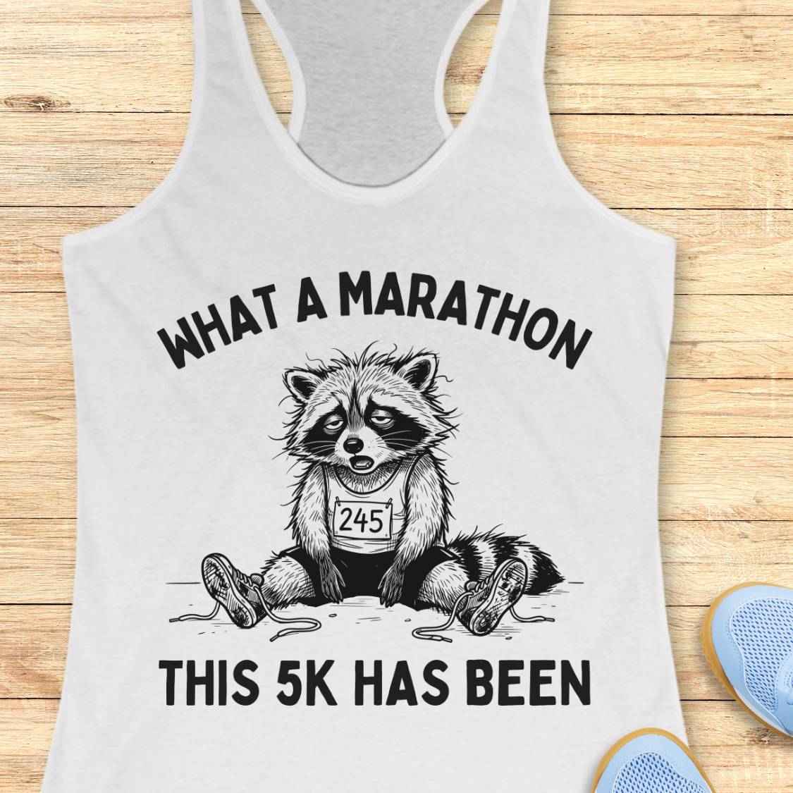 A white racerback tank top laid flat on a light wood surface, featuring a black graphic of a raccoon wearing a race bib and the text 'WHAT A MARATHON THIS 5K HAS BEEN.' Partial blue shoes are visible at the bottom right as styling props.