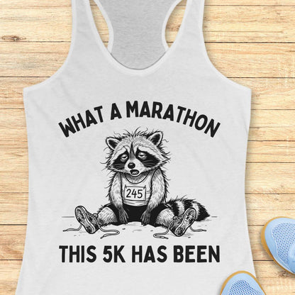 A white racerback tank top laid flat on a light wood surface, featuring a black graphic of a raccoon wearing a race bib and the text 'WHAT A MARATHON THIS 5K HAS BEEN.' Partial blue shoes are visible at the bottom right as styling props.