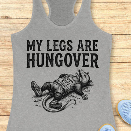 A heather gray racerback tank top laid flat on a light wood surface, featuring the black text 'MY LEGS ARE HUNGOVER' above a black illustration of a raccoon lying down with a race bib. Partial blue shoes are visible at the bottom right edge.