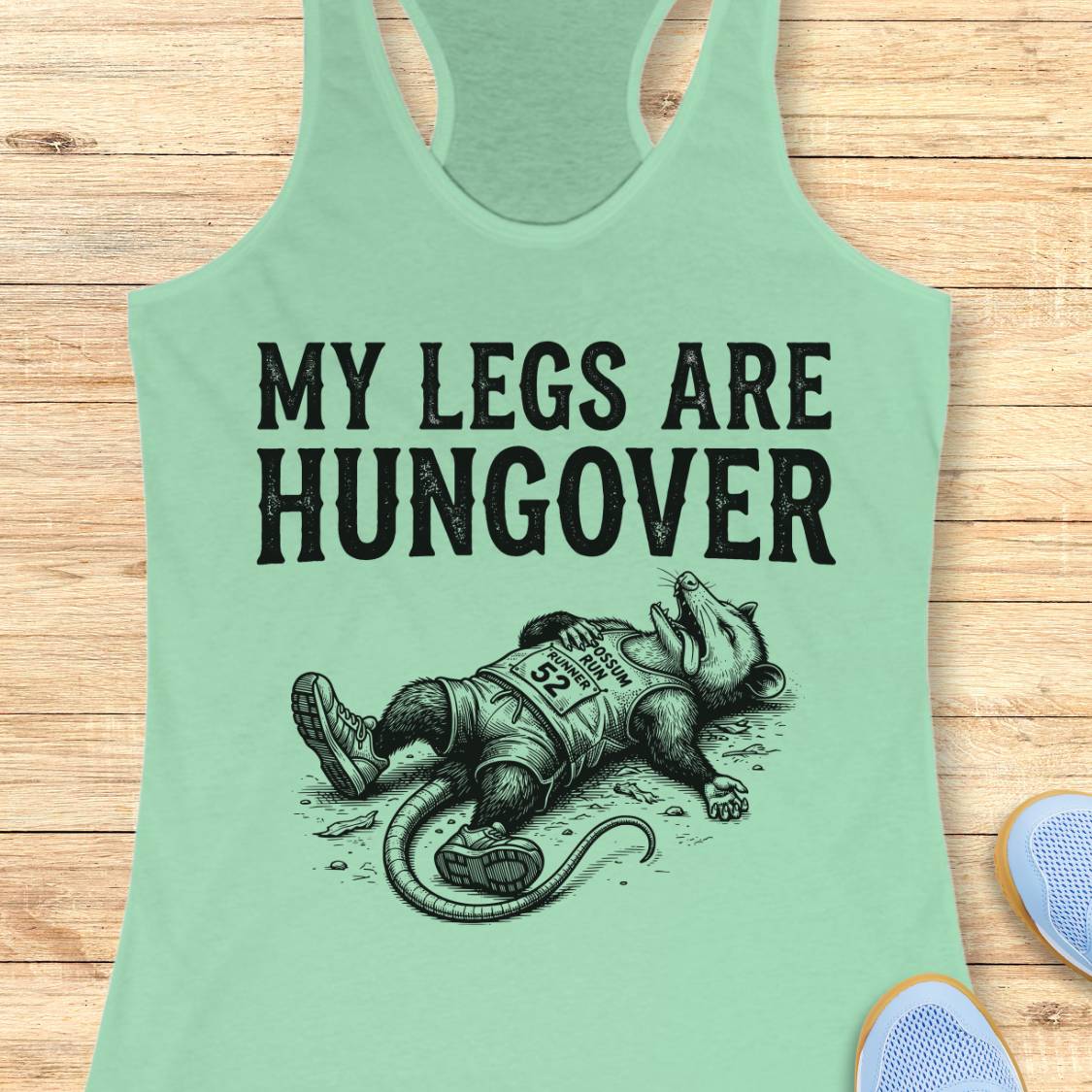 A mint green racerback tank top laid flat on a light wood background, featuring the black text 'MY LEGS ARE HUNGOVER' above a black illustration of a exhausted raccoon wearing a race bib. Partial blue sneakers are visible at the bottom right edge.