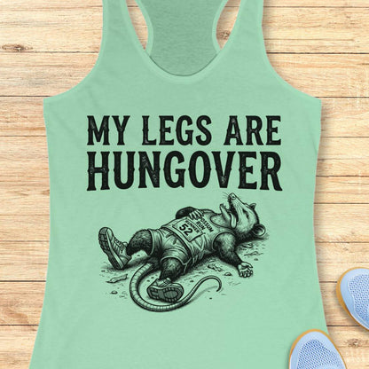 A mint green racerback tank top laid flat on a light wood background, featuring the black text 'MY LEGS ARE HUNGOVER' above a black illustration of a exhausted raccoon wearing a race bib. Partial blue sneakers are visible at the bottom right edge.