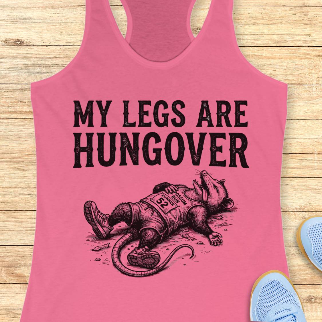 A pink racerback tank top laid flat on a light wood surface, featuring a black graphic of a exhausted racehorse wearing a bib numbered 92, with the text 'MY LEGS ARE HUNGOVER'.