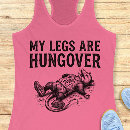 A pink racerback tank top laid flat on a light wood surface, featuring a black graphic of a exhausted racehorse wearing a bib numbered 92, with the text 'MY LEGS ARE HUNGOVER'.
