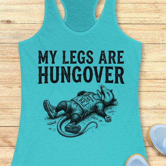 A turquoise racerback tank top laid flat on a light wood surface, featuring the black printed text 'MY LEGS ARE HUNGOVER' above an illustration of a person lying down in athletic wear. Part of a white shoe is visible at the bottom right.
