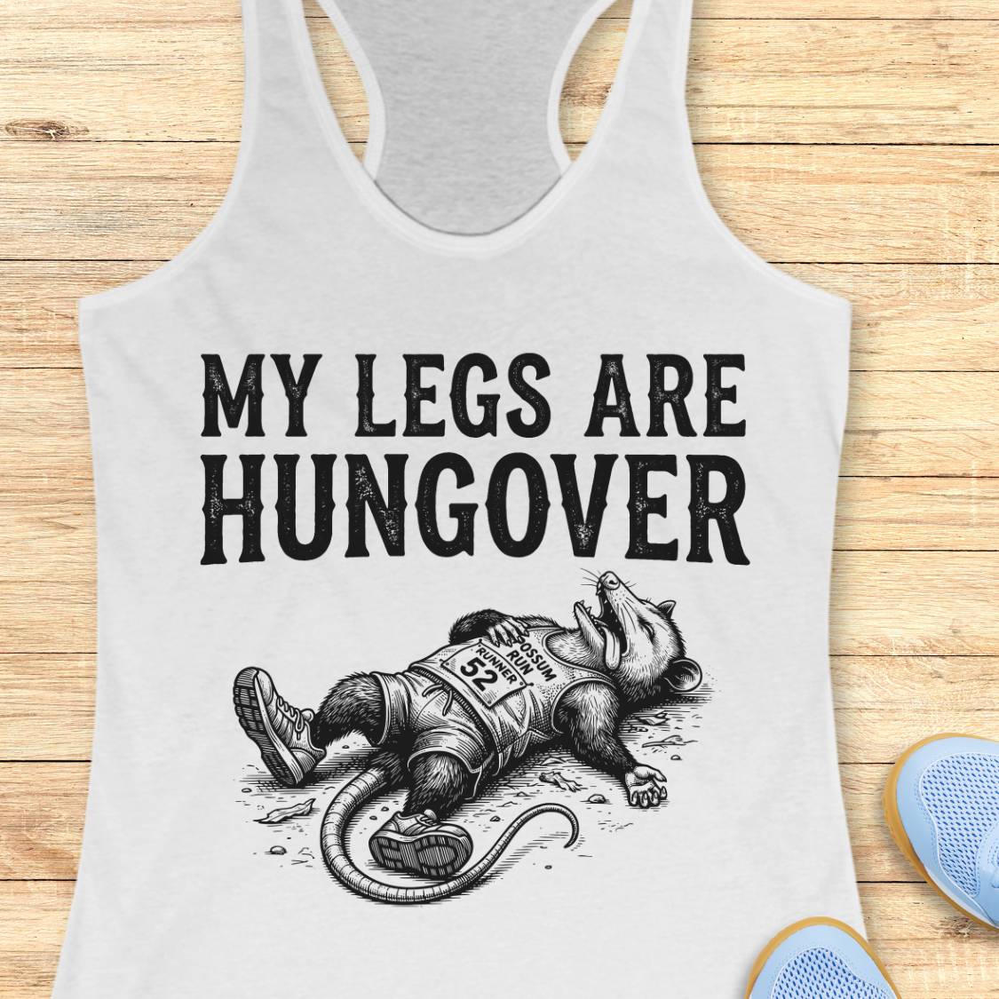 A white racerback tank top laid flat on a light wood plank background, featuring a black graphic print that reads 'MY LEGS ARE HUNGOVER' above an illustration of a exhausted raccoon wearing a race bib. Partial blue shoes are visible at the bottom right as styling props.