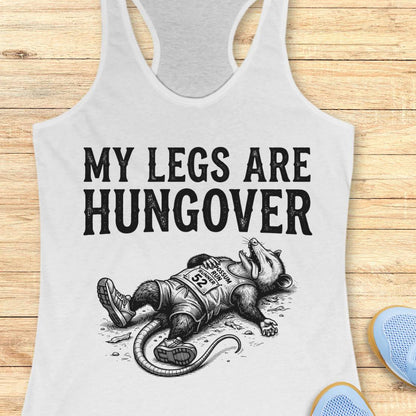 A white racerback tank top laid flat on a light wood plank background, featuring a black graphic print that reads 'MY LEGS ARE HUNGOVER' above an illustration of a exhausted raccoon wearing a race bib. Partial blue shoes are visible at the bottom right as styling props.